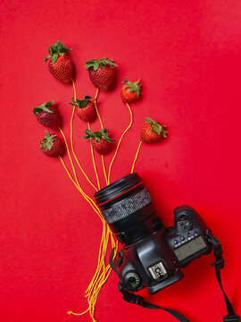 A camera sits with strawberries tied on strings floating above it like balloons, set against a bold red background in a creative concept