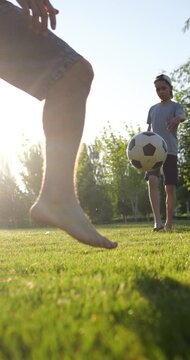 Barefoot boy passes football with father in park at sunset. Child practices soccer skills on grass while only legs of parent are visible in vertical video. Father and son playing football.