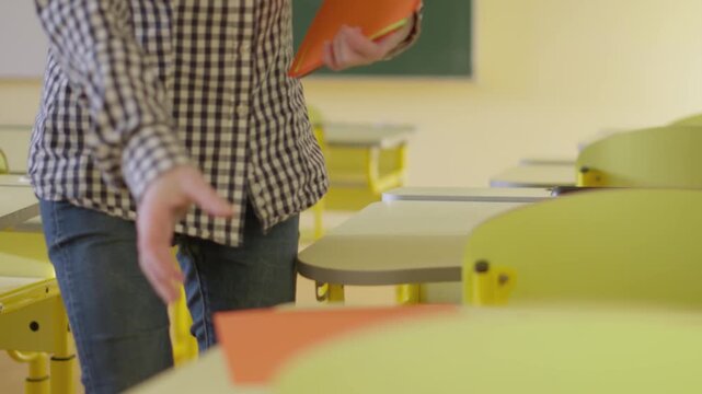 Close up of hands placing orange folders on modern yellow desks in a well lit classroom. The scene captures preparation, organization, and a welcoming educational environment