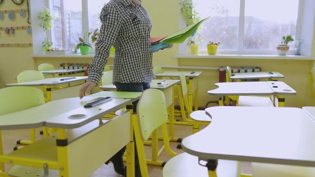 Contemporary classroom scene featuring empty desks, vibrant yellow furniture, and a teacher preparing for a lesson. Natural light and greenery create a welcoming learning space