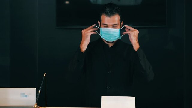 Handsome man putting on protective medical face mask and crossing arms, looking at camera on dark background. Concept of health protection, virus prevention, safety measures and new normal lifestyle