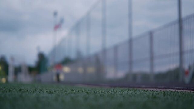 Cinematic close-up of artificial turf with a blurred athlete walking beside a football field at dusk. Shallow depth of field creates a dramatic sports and training atmosphere.

