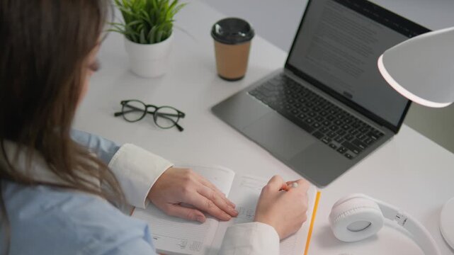 Young woman writes in notebook beside open laptop, headphones, coffee cup, creating productive study environment. Home office setup with green plant, glasses adds touch comfort, style