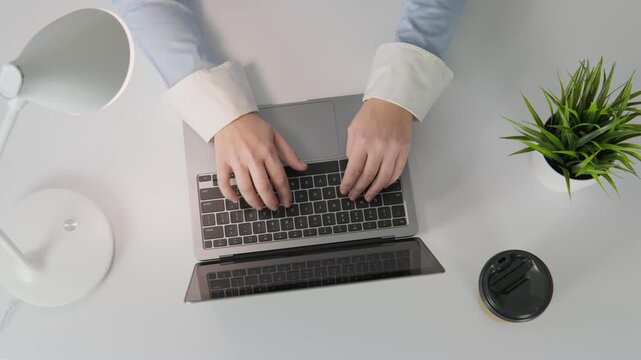 Hands on a laptop keyboard, surrounded by a coffee cup, a potted plant, and a desk lamp, create a clean and motivating workspace. Perfect for themes of remote work, freelancing, and productivity