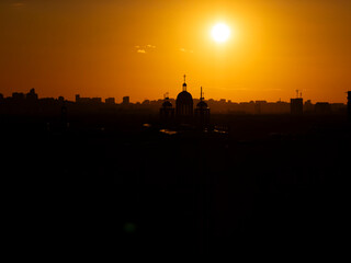 Obraz premium Urban cityscape showing building silhouettes and church domes against a bright orange sky during a vibrant sunset, representing tranquility and the beauty of evening light over a developing city