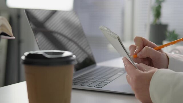 Person takes handwritten notes in a spiral notebook next to an open laptop, emphasizing multitasking and organization. Coffee and plants add a trendy, inspiring office vibe