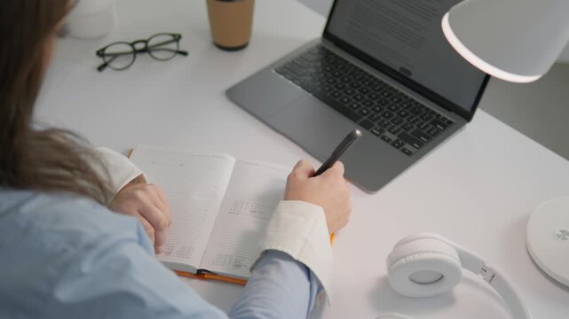 Young woman taking notes while studying online at a clean white desk with a laptop and headphones. Cozy workspace setup with soft lighting and minimalistic design for remote learning