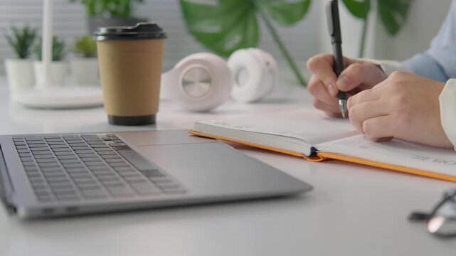 Contemporary office scene featuring a laptop, notebook, and takeaway coffee cup on a clean desk. Hands are writing in a notebook, emphasizing productivity and remote work trends