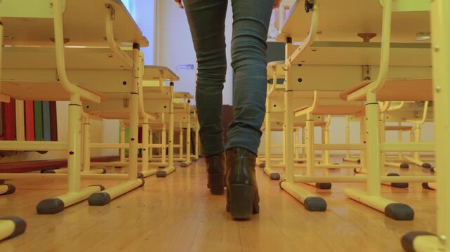 The image shows a rear view of an individual walking through a vacant classroom with neatly arranged desks and chairs. Warm lighting and clean floors create a welcoming academic atmosphere
