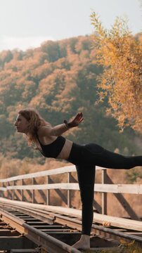 A fit woman performs a yoga pose on abandoned train tracks in a scenic autumn landscape. The golden foliage and serene setting highlight her focus, balance, and connection to nature