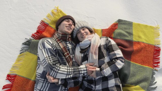 A cheerful couple relaxes on a vibrant blanket atop fresh snow, dressed in winter attire and sharing a joyful embrace. Their laughter and closeness capture the essence of winter romance