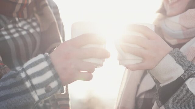 Close up of hands holding mugs, wrapped in scarves and jackets, sharing a hot beverage outside. The sunlight creates a gentle, inviting atmosphere, perfect for winter relaxation