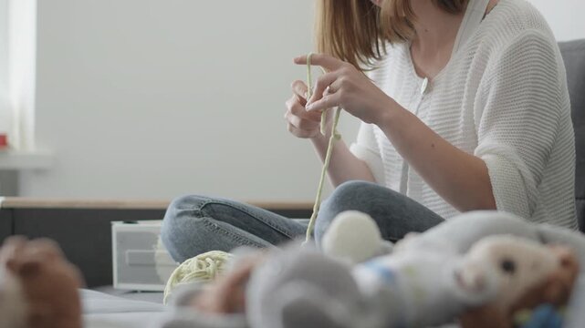 A woman in casual clothes is knitting on a comfortable sofa, surrounded by handmade toys and yarn. The scene captures a peaceful, creative moment in a stylish, minimalist home