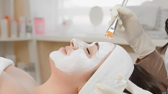 Aesthetician in gloves applies advanced facial treatment to a client with a white mask in a bright, clean spa environment. The scene highlights modern skincare and wellness trends