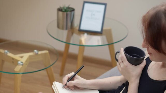 Young woman enjoys a hot drink while journaling in a stylish, light filled room featuring glass tables and wooden legs. The scene reflects relaxation, mindfulness, and contemporary home decor trends