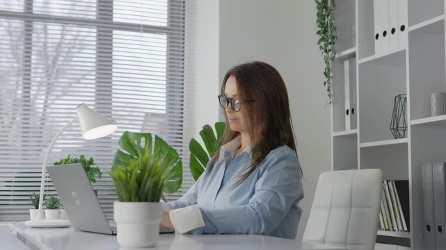 Young woman in glasses types on laptop at contemporary office table surrounded by indoor plants. Productive workplace scene with natural light, minimalistic decor, and relaxed professional atmosphere