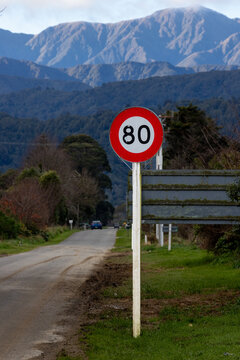 An 80 km/h speed limit sign stands along a quiet, scenic rural road in the South Island of New Zealand, framed by beautiful mountain peaks.