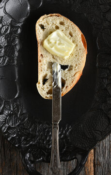 Bread still life. A fresh baked slice of bread on a black platter with a knife and smear of butter on the slice.