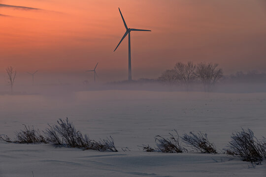 Wind turbines in a snowy field covered in thick morning fog at sunrise along Highway 20, Niagara Peninsula, Ontario, Canada.