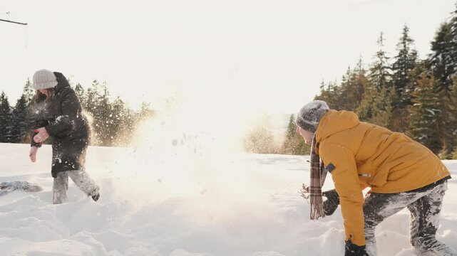 Friends dressed in warm winter clothing throw snowballs at each other in snowy field, with sunlight illuminating scene. Joyful moment captures essence outdoor winter fun, friendship