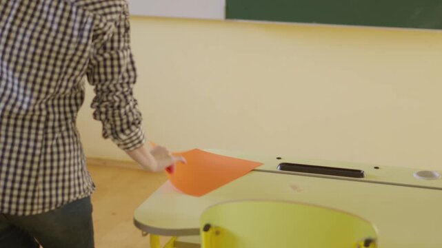 A teacher stands in a modern classroom, holding colored sheets while preparing for class activities. The room features yellow furniture and a welcoming atmosphere for students