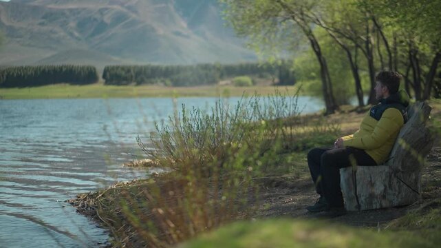 Man wearing a yellow jacket sitting on a wooden bench overlooking a calm lake and snow-capped mountain peaks at Laguna La Zeta, Esquel, Chubut, Argentina.