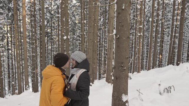 Two people dressed in winter clothing enjoy a quiet moment in a serene, snow covered forest. The tall trees and untouched snow create a magical, tranquil winter atmosphere