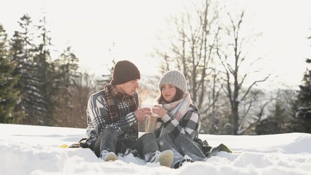 Young man and woman in knit hats and plaid shirts relax on a blanket in the snow, sipping from mugs. The winter forest background and bright sunlight evoke comfort and togetherness