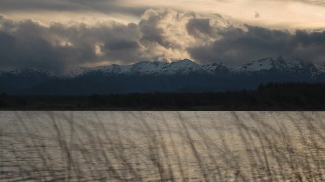 Dramatic view of snow-capped mountains reflecting over the dark, rippling waters of a lake during a cloudy sunset.