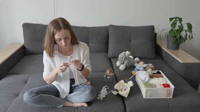 Focused female artisan creates knitted toys in a bright living room, surrounded by handmade dolls and craft supplies. Creative hobby scene highlights slow living and mindfulness