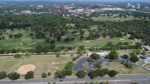 Lions Field baseball diamonds rest across roadway from Brackenridge Park Golf Course. Asphalt parking lots border manicured turf facilities under bright afternoon daylight, San Antonio, Texas