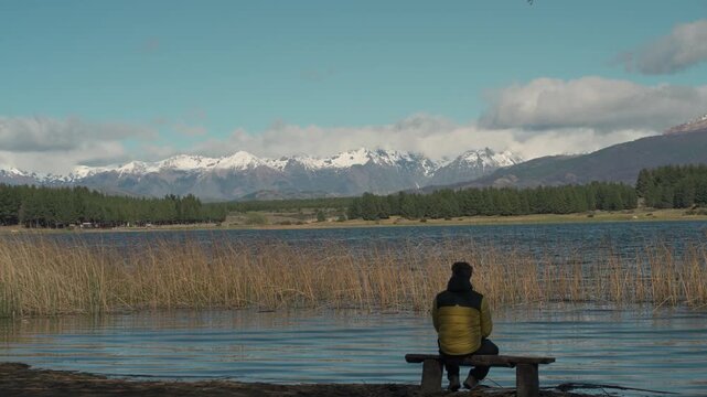 Man wearing a jacket sitting and contemplating the scenic view of Laguna La Zeta, surrounded by dense pine forests and snow-capped mountains in Esquel, Patagonia, Argentina.