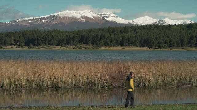 Man in a yellow jacket walking along a trail surrounded by lush vegetation and trees next to the calm waters of Laguna La Zeta, Esquel, Chubut, Argentina.