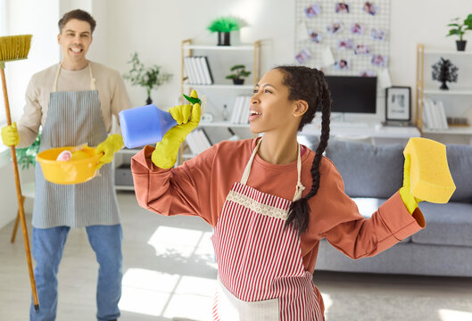 Couple cleaning home livingroom singing broom dustpan teamwork. Smiling woman sings into spray bottle as man holds broom and dustpan, enjoying cheerful housework. Joyful teamwork and tidy home.