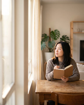 Asian woman pausing to reflect while writing in a journal at home