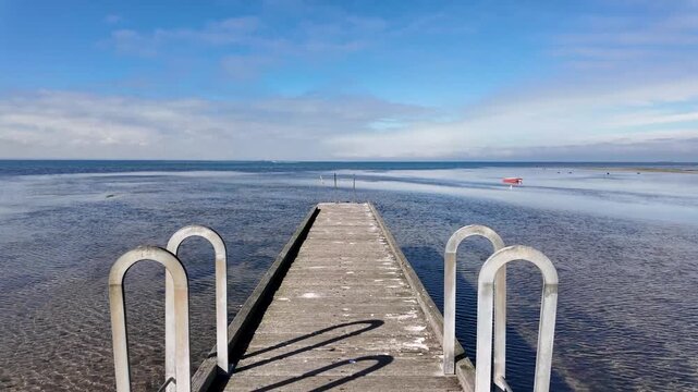 POV perspective walking along the wooden deck of Seaholme Jetty, extending out towards Port Phillip Bay in Melbourne, Australia, with expansive shallow water, exposed sandbars at low tide