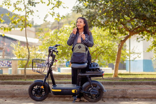 Young Black woman applying lip gloss beside her electric bicycle, taking a moment for herself during her daily routine.