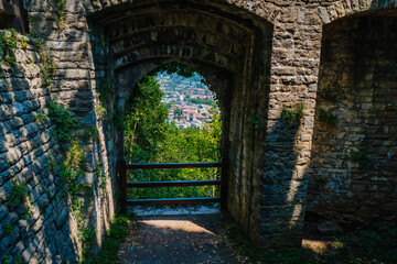 Obraz premium Stone archway leading to a scenic view of Bergamo city from Rocca di Bergamo park, surrounded by greenery and historic architecture