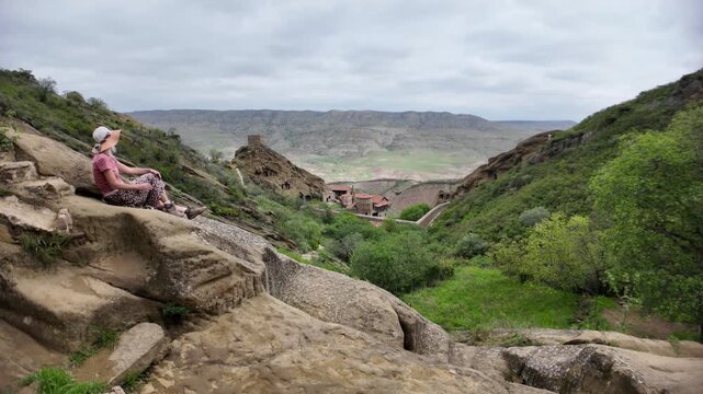 Woman resting on ancient rock steps, enjoying the panoramic view of the historic David Gareji rock-hewn monastery complex nestled in the green valley landscape of Georgia's Middle Ages heritage site