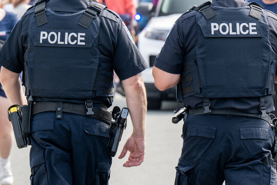 Two police officers wearing blue uniforms with white lettering on their backs. The cops are wearing guns, handcuffs, tasers, protective bulletproof vests, tactical belts, and narcotic testing kits.
