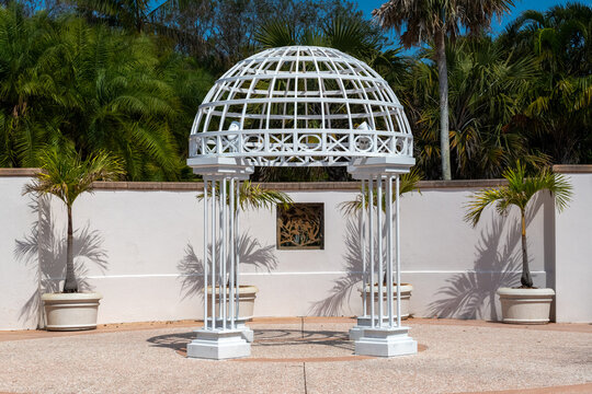A large white metal gothic-style archway dome in an enclosed outdoor garden. The wedding structure has four heavy-duty iron pillars. The background is lush green trees and shrubs. The ground is a bric