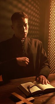 Priest praying with Holy Bible and rosary at wooden table in confessional booth