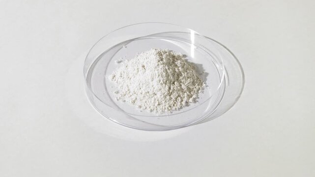 Vertical jib shot of white fine powder piled in a plastic petri dish on a pale gray canvas lit by moving light creating dynamic shadows | Titanium dioxide cosmetic ingredient concept