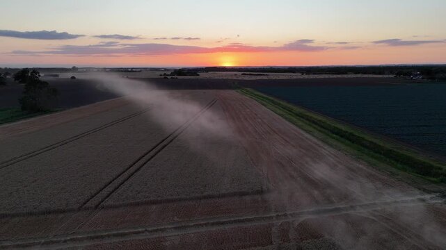 Aerial view of dust plume over farmland at sunrise. Sun glows low, casting warm hues across horizon. Furrows stretch through dry earth, contrasting with green fields