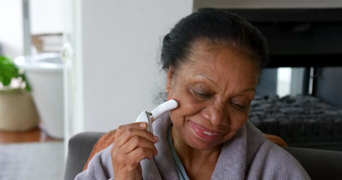 Senior African American woman in gray bathrobe doing skincare on sofa, pressing wand pulsing LED