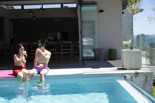 Asian couple sitting with feet in pool on rooftop deck in swimwear holding drinks, copy space