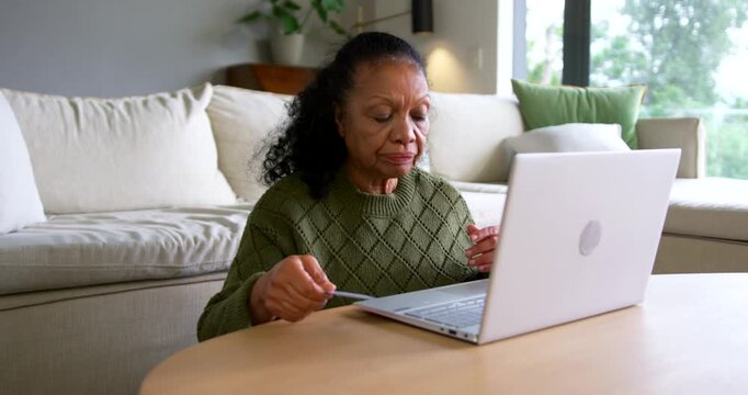 Senior African American woman holding dark card, inspecting then typing card info on laptop at home