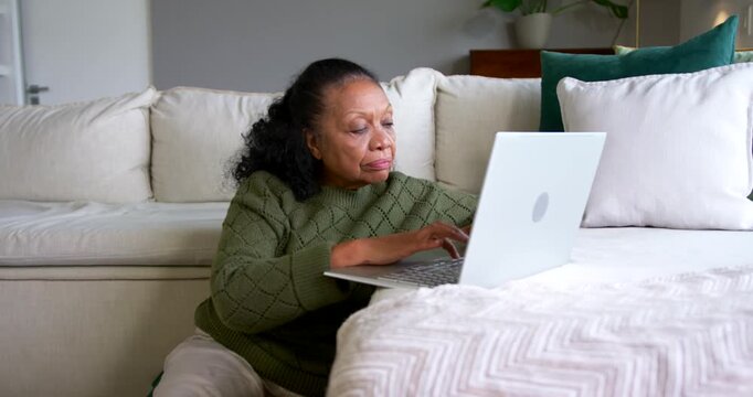 Senior African American woman placing laptop on ottoman at home, typing to finish emails in sweater