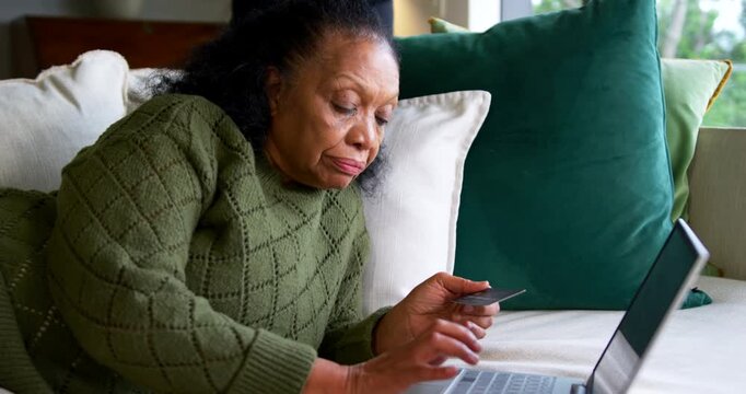 Senior African American woman lounging on couch in green sweater, typing on laptop with card buying