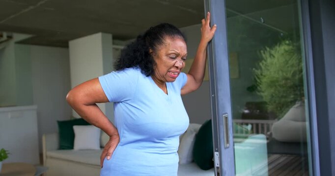 Senior African American woman feeling sudden back pain, leaning on sliding glass door frame
